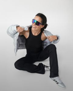 Una donna è seduta sul pavimento e si infila sorridendo una felpa grigia sopra la testa. Indossa un top nero senza maniche, pantaloni da jogging grigi, calzini bianchi e scarpe da ginnastica grigie su uno sfondo semplice grigio chiaro.