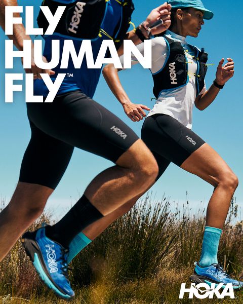 Due corridori, un uomo e una donna, corrono su un sentiero di montagna roccioso, circondati da una vegetazione lussureggiante e alberi alti. Entrambi indossano abbigliamento da corsa e zaini da idratazione. In alto spicca chiaramente la parola "HOKA". Sullo sfondo si ergono montagne innevate.