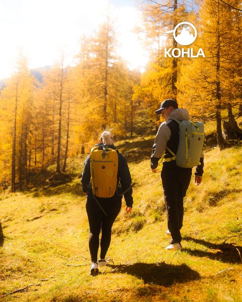 Due escursionisti con zaini camminano attraverso una foresta soleggiata con alberi autunnali dorati. La luce del sole filtra tra i rami, proiettando una luce calda. Il logo Kohla è visibile nell'angolo in alto a destra.