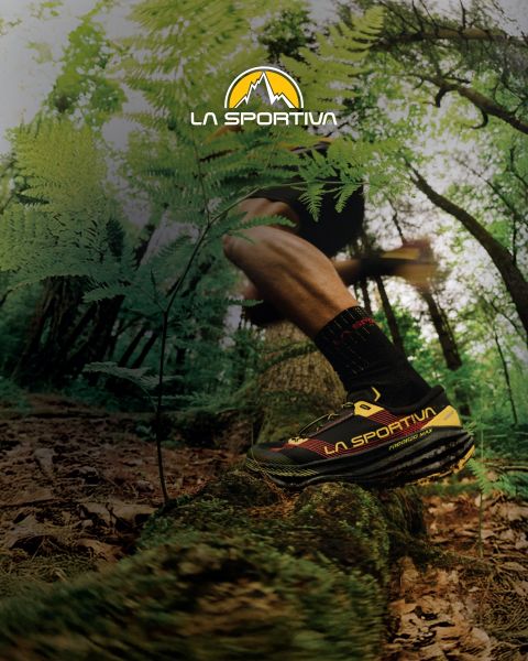 Quattro persone con attrezzatura da outdoor e materassini da boulder sulle spalle percorrono un sentiero di montagna erboso. Sopra di loro, nel cielo, si vede il logo di La Sportiva. Sullo sfondo si intravedono aspre cime montuose.