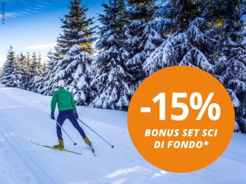 Una persona pratica sci di fondo su una pista innevata fiancheggiata da pini sotto un cielo azzurro. Un cerchio arancione indica: "-15% BONUS SET SCI DI FONDO*".