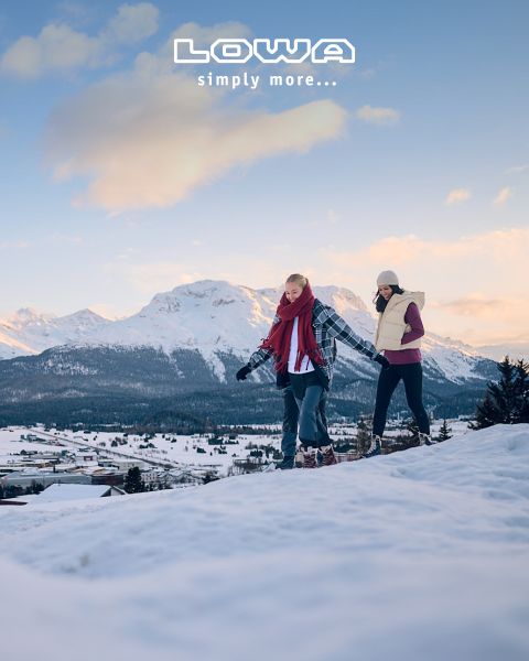 Tre persone fanno escursioni con bastoncini da trekking e attrezzatura da esterno su un pittoresco sentiero di montagna. Sullo sfondo si vedono alberi e montagne. Il cielo è nuvoloso. In alto c'è la scritta "LOWA simply more…".
