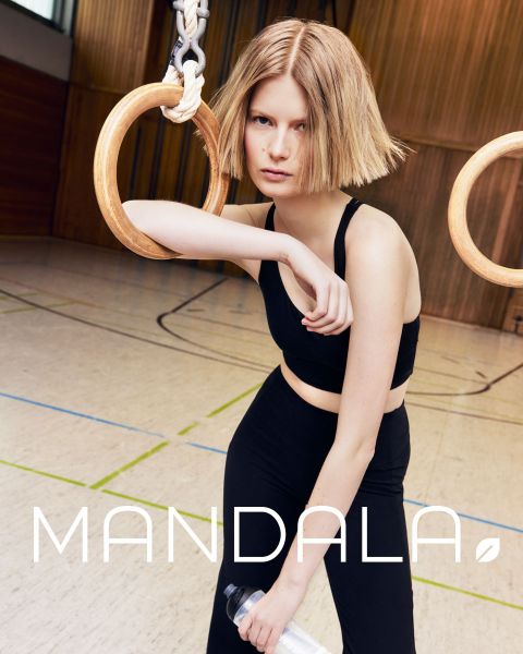 Donna con cappello e canotta nera guarda direttamente la fotocamera. I suoi capelli sono ondulati e sciolti. L'immagine è in bianco e nero e in basso, a grandi lettere, c'è la parola "MANDALA".