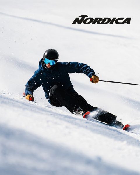 Due sciatori con equipaggiamento protettivo - uno in blu, l'altro in abiti scuri - scendono una montagna innevata. Il logo "Nordica" è visibile in alto. Entrambi sono inclinati, dimostrando velocità e agilità in una giornata di sole.