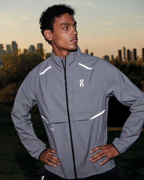 Una persona corre all'aperto. Indossa una giacca a vento beige-grigia, pantaloncini grigio chiaro e scarpe da corsa bianche. Lo sfondo è un cielo sereno e una superficie piana. Il logo "On" è visibile nell'angolo in alto a destra.