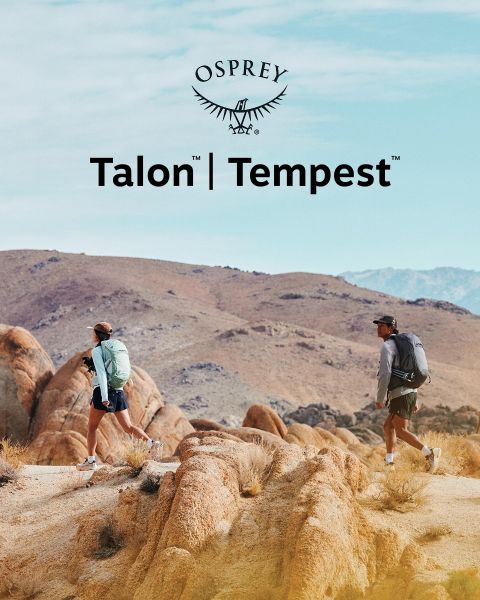 Una persona scende un terreno roccioso con uno zaino verde, un cappello nero e abiti scuri. La luce del sole illumina il paesaggio. In primo piano si vede il logo Osprey.