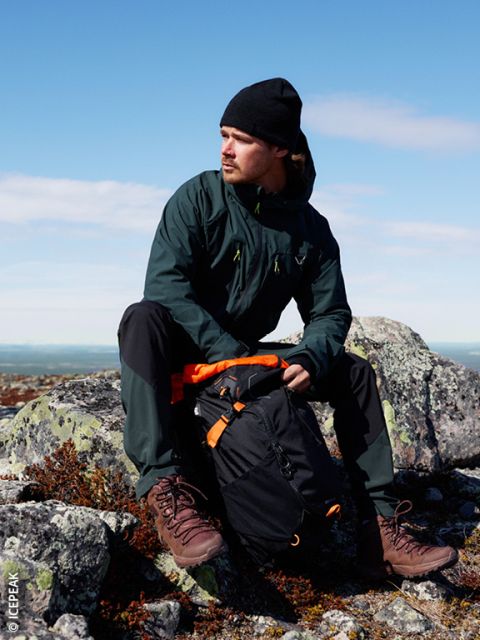 Un uomo con giacca scura, berretto nero e scarponcini da trekking è seduto su un terreno roccioso all'aperto e indossa uno zaino nero e arancione. Il cielo è limpido e blu sullo sfondo.