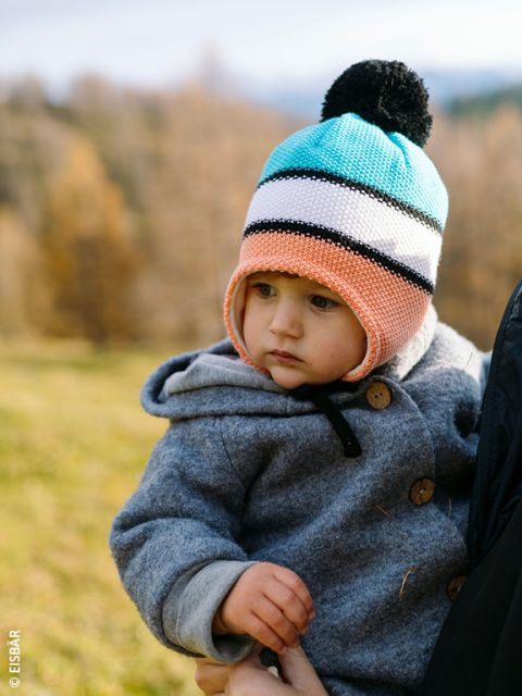 Un bambino piccolo che indossa un berretto di maglia colorato e un cappotto grigio viene tenuto in braccio da un adulto all'aperto, con uno sfondo sfocato di alberi autunnali ed erba.