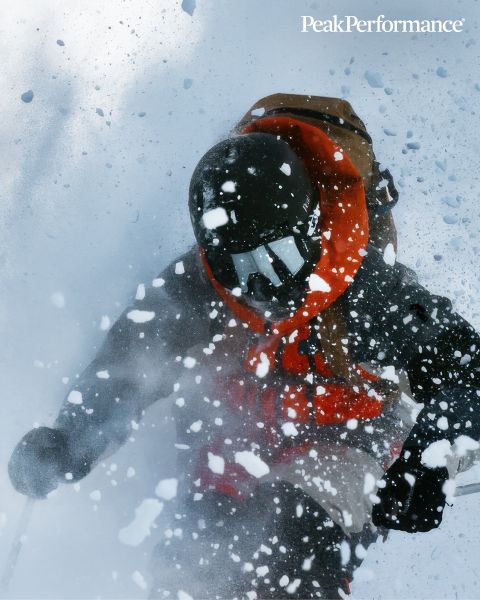 Una persona salta tra grandi rocce in una regione montuosa sotto un cielo azzurro brillante. Indossa abbigliamento sportivo, tra cui una giacca e pantaloncini. In alto si vede la scritta "Peak Performance".