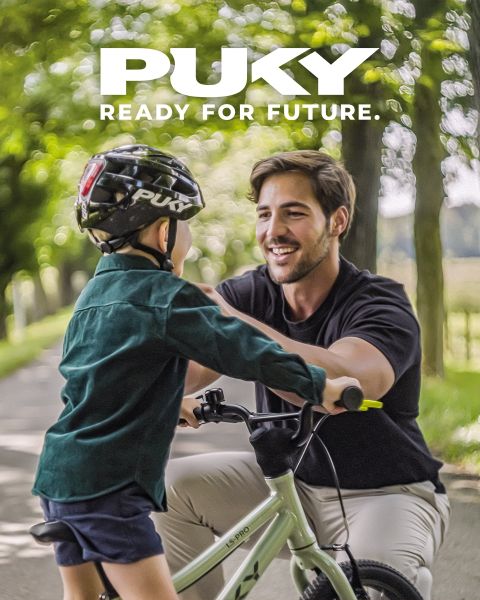 Una giovane ragazza con casco blu e maglietta a righe va in bicicletta azzurra in una giornata di sole. Sorridendo, passa accanto a graffiti colorati in un ambiente urbano. Il logo PUKY e lo slogan "Ready for life" sono visibili in alto a sinistra.