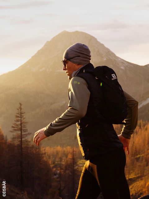 Una donna che indossa una giacca e guanti rosa-grigi corre in un paesaggio montano innevato e porta due bottiglie d'acqua nelle tasche anteriori del suo gilet da corsa.