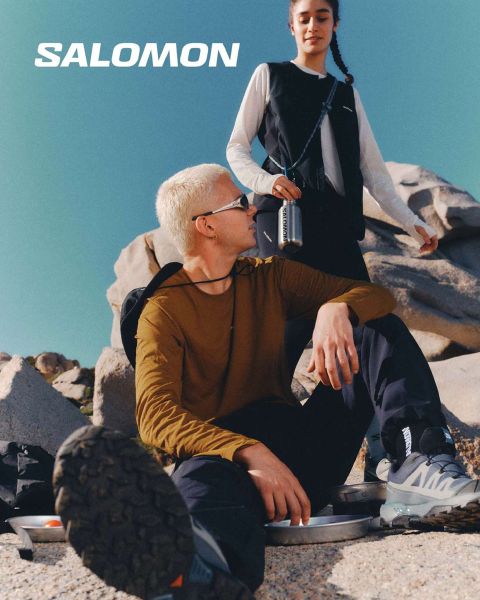 Primo piano di una persona che indossa scarpe Salomon e calze Salomon con gradiente blu. Si trova su un prato con un pittoresco paesaggio montano sullo sfondo. Un escursionista in abbigliamento outdoor si trova più lontano, anch'esso sul prato.