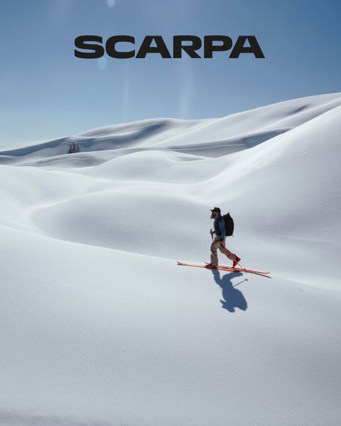 Una donna e un uomo sono seduti all'aperto su grandi rocce. Indossano abbigliamento casual e occhiali da sole. Sono circondati da verde e rocce, il cielo è leggermente nuvoloso. Nella parte superiore dell'immagine si vede la parola "SCARPA".