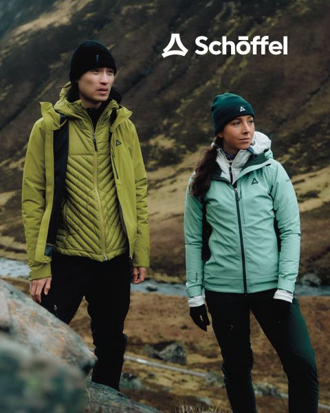 Due persone con abbigliamento da outdoor camminano su una collina erbosa con montagne e nuvole sullo sfondo. Sopra di loro c'è il logo Schöffel, sotto una linea ondulata verde decorativa.