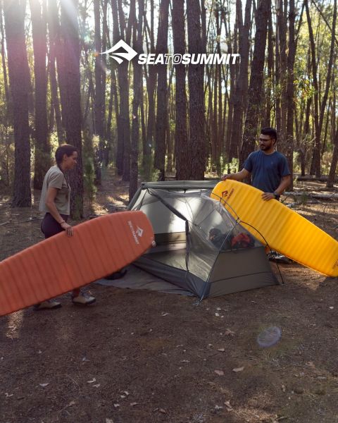 Una persona è sdraiata su un materassino isolante, avvolta in un sacco a pelo giallo, e osserva un'alba su una scogliera costiera. Attrezzatura da campeggio è accanto a lei, e le onde del mare si infrangono contro le rocce. Il logo "SEATOSUMMIT" si trova nell'angolo in basso a destra.