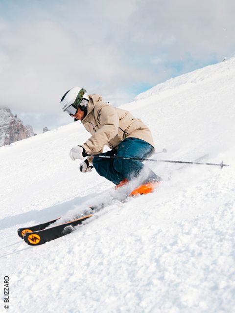 Uno sciatore con giacca beige, pantaloni blu e casco bianco scende lungo una pista innevata, affronta una curva stretta sollevando neve. Sullo sfondo si vedono le montagne.