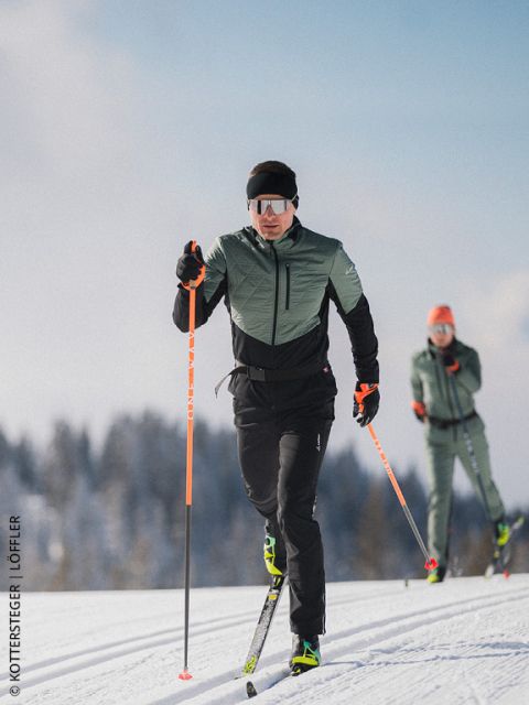 Due sciatori di fondo su una pista innevata, con attrezzatura da sport invernali e occhiali da sole, con alberi e cielo azzurro sullo sfondo.