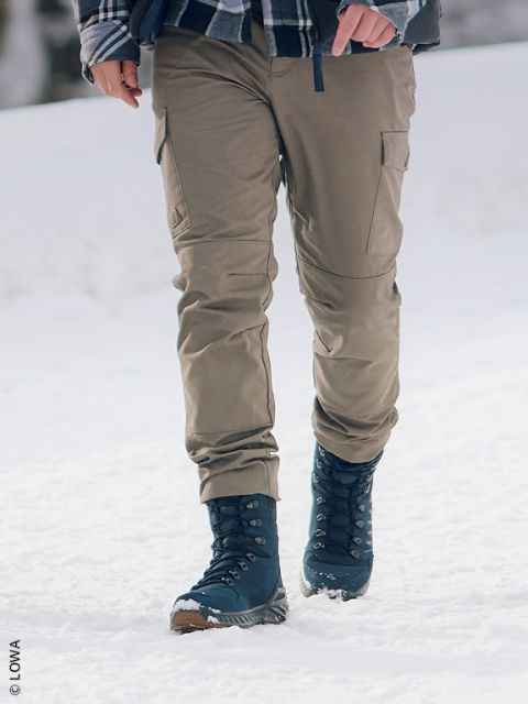 Persona con pantaloni color kaki e stivali invernali blu navy che cammina sulla neve, visibili solo le gambe e una parte del busto.