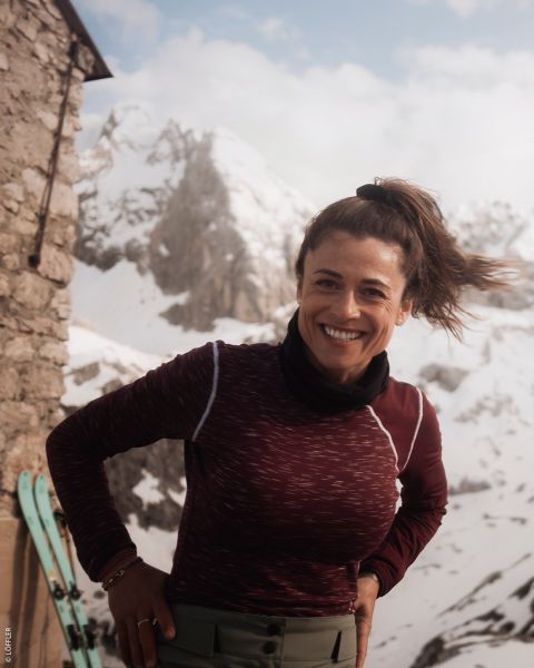 Una donna sorridente con un top marrone castagna è in piedi all'aperto vicino a un edificio in pietra con montagne innevate e sci sullo sfondo.