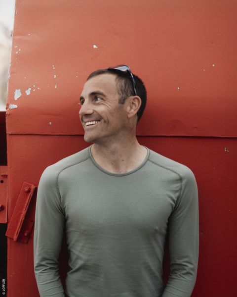 Un uomo sorridente con una camicia verde è in piedi davanti a una grande struttura metallica rossa e guarda di lato con un paio di occhiali da sole sulla testa.