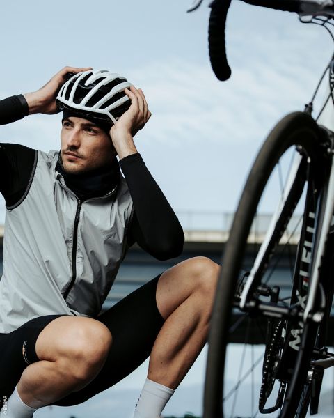 Un ciclista con casco e abbigliamento da ciclista è inginocchiato accanto a una bicicletta e si sistema il casco, con un ponte e il cielo sullo sfondo.