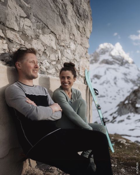 Due persone in abbigliamento invernale sono sedute sul sedile posteriore di un veicolo parcheggiato su una strada innevata. Entrambe sorridono. La persona a sinistra indossa un berretto nero e una felpa, mentre la persona a destra indossa un maglione fantasia, un casco e degli occhiali protettivi. Testo nell'immagine.