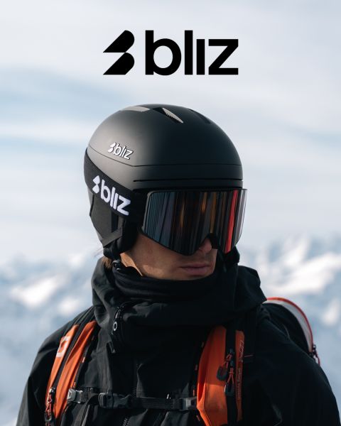 Una persona che indossa un casco da sci nero e una maschera da sci a specchio, entrambi con la scritta "Bliz", è in piedi all'aperto con montagne innevate sullo sfondo. Il logo Bliz è ben visibile nella parte superiore dell'immagine.