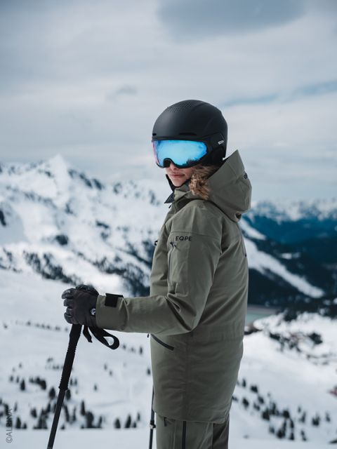 Uno sciatore in giacca verde e casco nero si trova su un pendio montuoso innevato, tiene i bastoncini da sci e si guarda indietro, con cime sullo sfondo.