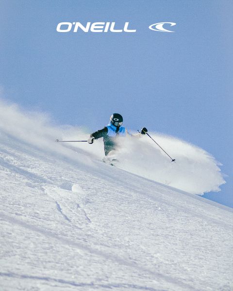 Una donna con i capelli lunghi si trova in un paesaggio desertico sotto un cielo blu con nuvole sparse. Indossa una canotta nera O'Neill e si sporge leggermente in avanti. Sul lato destro dell'immagine si vede il logo O'Neill.
