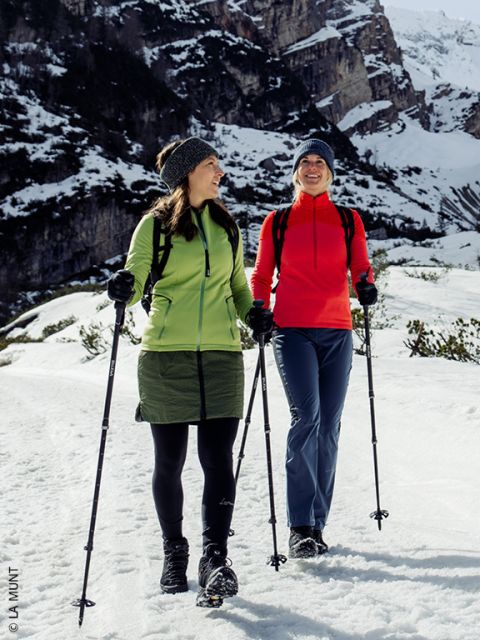 Due donne in abbigliamento invernale camminano con bastoncini su un sentiero innevato e si sorridono sullo sfondo di montagne innevate.