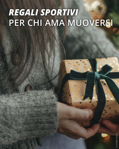 Persona con un maglione grigio che tiene in mano una confezione regalo avvolta in carta dorata con un nastro verde scuro. Il testo in italiano recita: "Regali sportivi per chi ama muoversi."