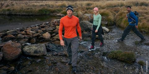 Tre persone con attrezzatura da esterno attraversano un ruscello roccioso in una zona montuosa e erbosa, con una persona che precede e le altre che seguono.