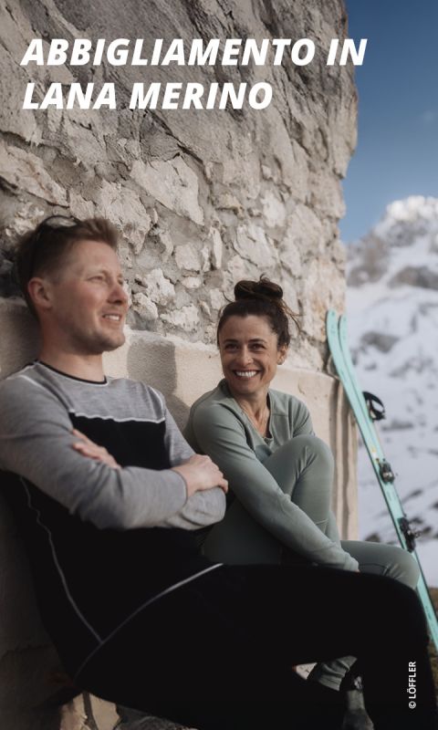 Un uomo sorridente e una donna sorridente in abbigliamento da sci sono seduti all'aperto su un muro di pietra, con gli sci nelle vicinanze e montagne innevate sullo sfondo.