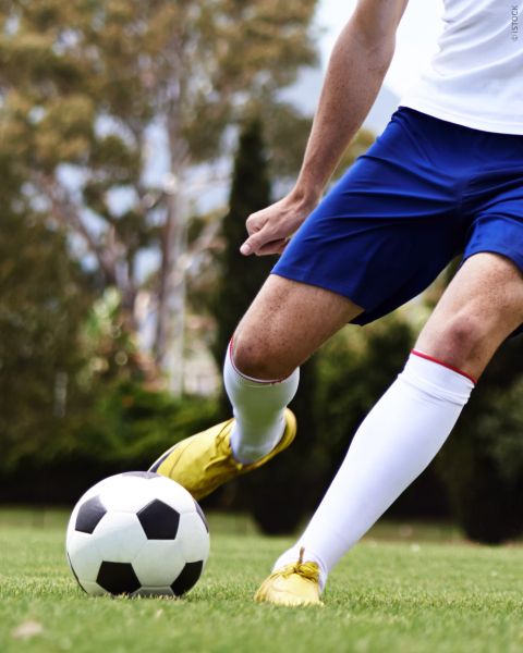 Un calciatore in pantaloncini blu e scarpe gialle che calcia un pallone da calcio bianco e nero su un prato, con alberi sullo sfondo.