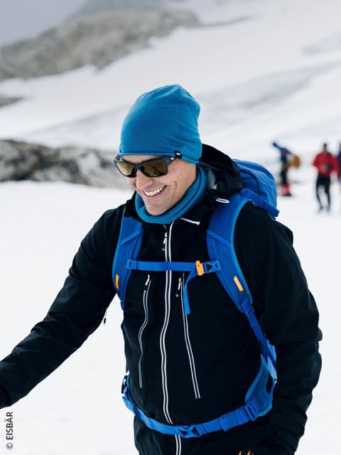 Una persona sorridente in abbigliamento invernale cammina in un paesaggio innevato, indossa occhiali da sole, un cappello blu e uno zaino blu, mentre sullo sfondo si vedono altre persone.