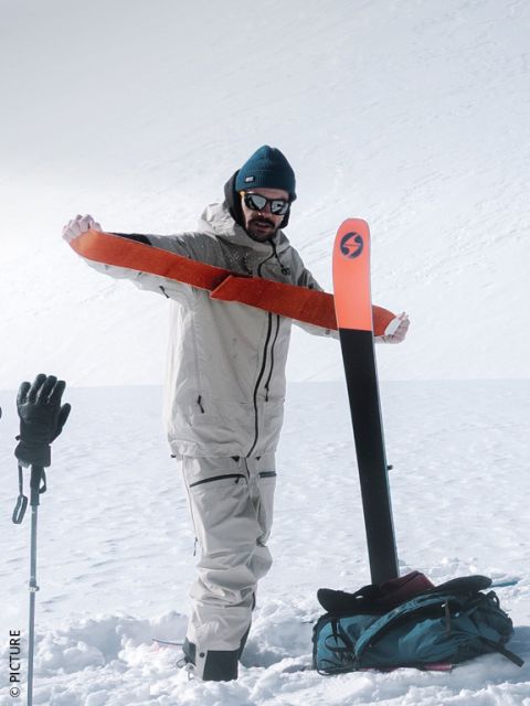 Una persona con equipaggiamento invernale tira una pelle di foca arancione su uno sci su una montagna innevata, mentre porta con sé bastoncini da sci e uno zaino.