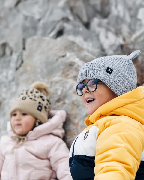 Due bambini piccoli con giacche invernali e berretti di lana sorridono seduti all'aperto, davanti a uno sfondo roccioso. Uno indossa il giallo, l'altro il rosa.