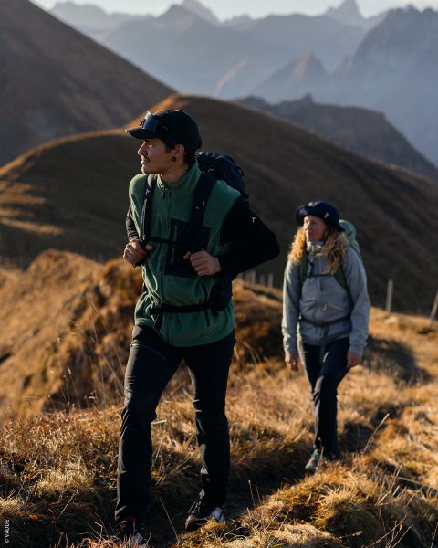 Due escursionisti con attrezzatura da esterno camminano su un sentiero di montagna soleggiato con cime frastagliate e colline erbose sullo sfondo.