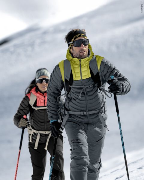 Due persone con equipaggiamento invernale camminano su un pendio montuoso innevato con bastoncini da sci, occhiali da sole e zaini sotto un cielo luminoso e nuvoloso.