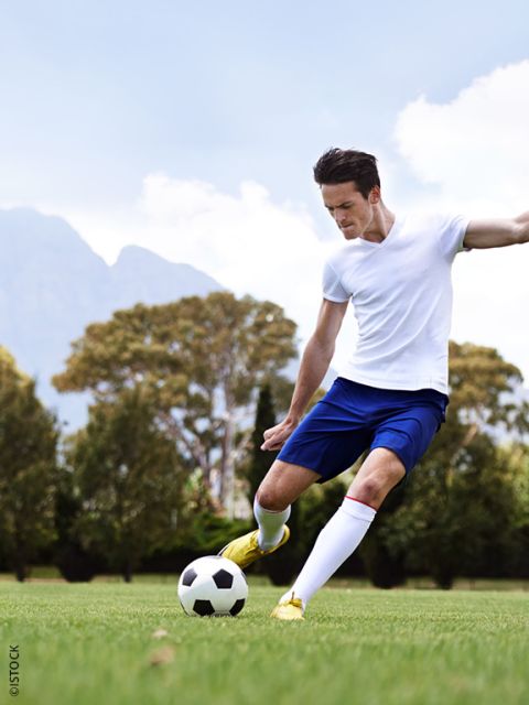 Un calciatore in una maglia bianca e pantaloncini blu calcia un pallone da calcio su un prato con alberi e montagne sullo sfondo.