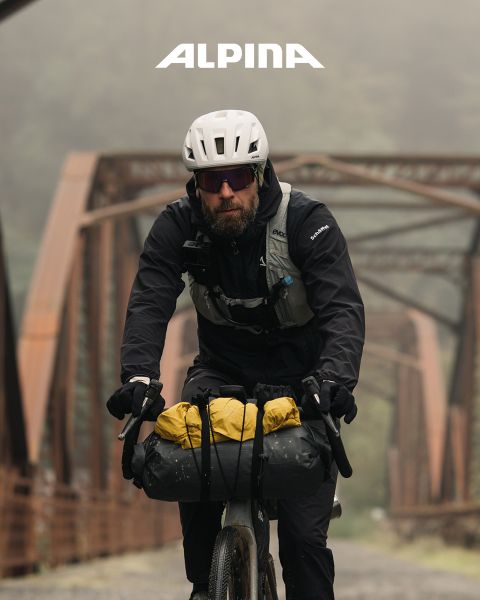 Un ciclista con casco e camicia bianca è in piedi con la sua bicicletta e guarda in lontananza al tramonto. Sullo sfondo si vedono alberi, colline e una linea elettrica. Sopra si vede la parola "ALPINA".