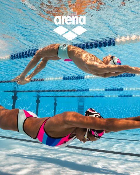Un nuotatore con cuffia nera e occhialini riflettenti scivola sott'acqua in una piscina delimitata da corsie. Il costume da bagno blu presenta decorazioni arancioni. L'acqua è limpida e sopra si vede il logo Arena.