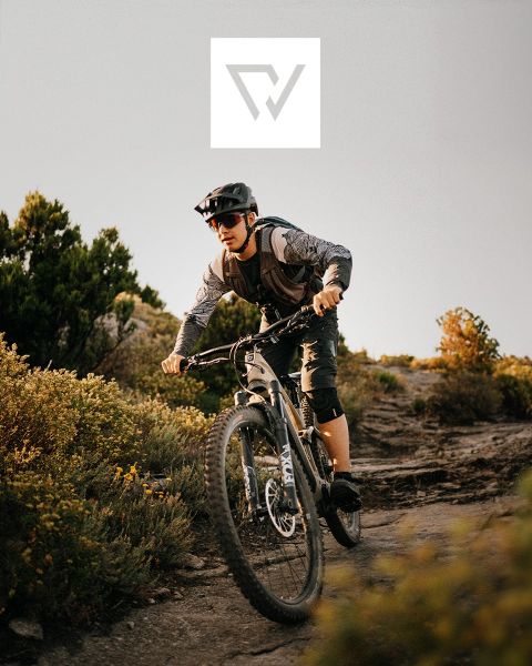 Due ciclisti stanno percorrendo un sentiero accidentato con mountain bike in una regione montuosa con cime innevate sullo sfondo. Indossano caschi e attrezzatura da outdoor. Nella parte superiore dell'immagine è visibile un logo.