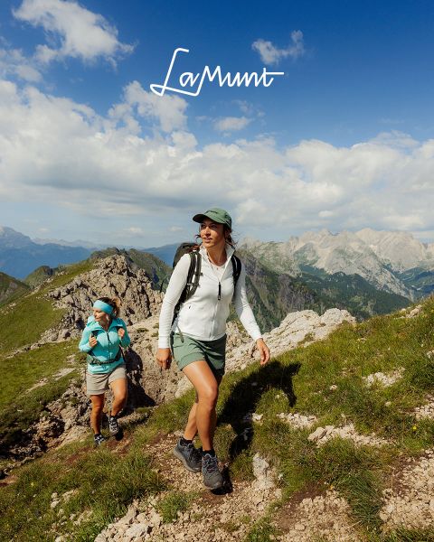 Tre persone in abbigliamento invernale sciano in un paesaggio montano innevato sotto un cielo limpido. Il testo recita: "LaMunt MOUNTAIN ME-TIME". Sullo sfondo si vedono cime innevate.