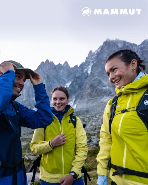 Due escursionisti con attrezzatura da outdoor attraversano un terreno roccioso sotto un cielo nuvoloso, con una grande montagna sullo sfondo. Entrambi indossano abbigliamento Mammut e il logo del marchio è visibile nella parte superiore dell'immagine.