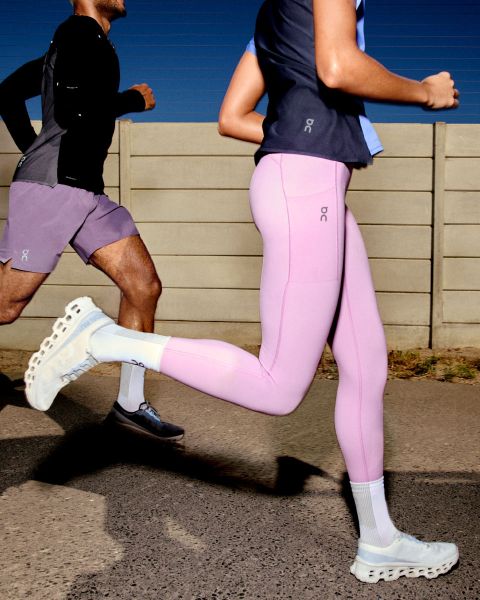 Una persona corre all'aperto. Indossa una giacca a vento beige-grigia, pantaloncini grigio chiaro e scarpe da corsa bianche. Lo sfondo è un cielo sereno e una superficie piana. Il logo "On" è visibile nell'angolo in alto a destra.