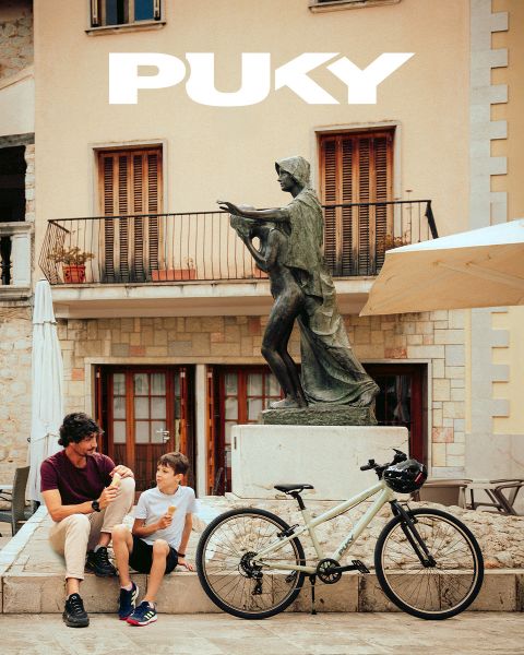 Una giovane ragazza con casco blu e maglietta a righe va in bicicletta azzurra in una giornata di sole. Sorridendo, passa accanto a graffiti colorati in un ambiente urbano. Il logo PUKY e lo slogan "Ready for life" sono visibili in alto a sinistra.