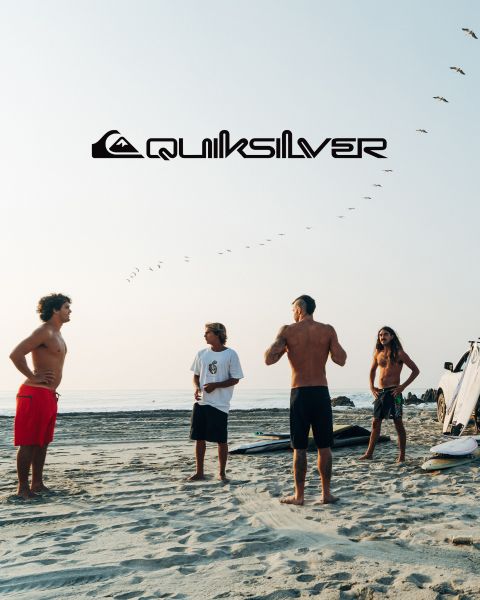 Un surfista cavalca una grande onda turchese, incarnando equilibrio e abilità. Indossa pantaloncini da bagno scuri e l'onda si increspa sopra di lui. Sopra di lui si vede il logo Quiksilver.