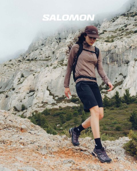 Primo piano di una persona che indossa scarpe Salomon e calze Salomon con gradiente blu. Si trova su un prato con un pittoresco paesaggio montano sullo sfondo. Un escursionista in abbigliamento outdoor si trova più lontano, anch'esso sul prato.