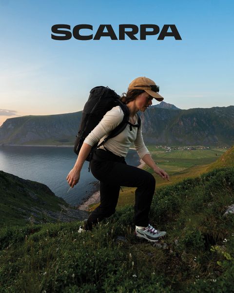 Una donna e un uomo sono seduti all'aperto su grandi rocce. Indossano abbigliamento casual e occhiali da sole. Sono circondati da verde e rocce, il cielo è leggermente nuvoloso. Nella parte superiore dell'immagine si vede la parola "SCARPA".